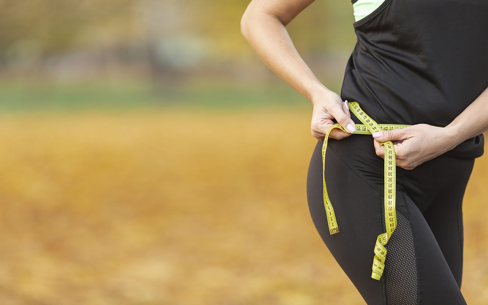 A woman in athletic wear measuring her waist with a tape measure outdoors