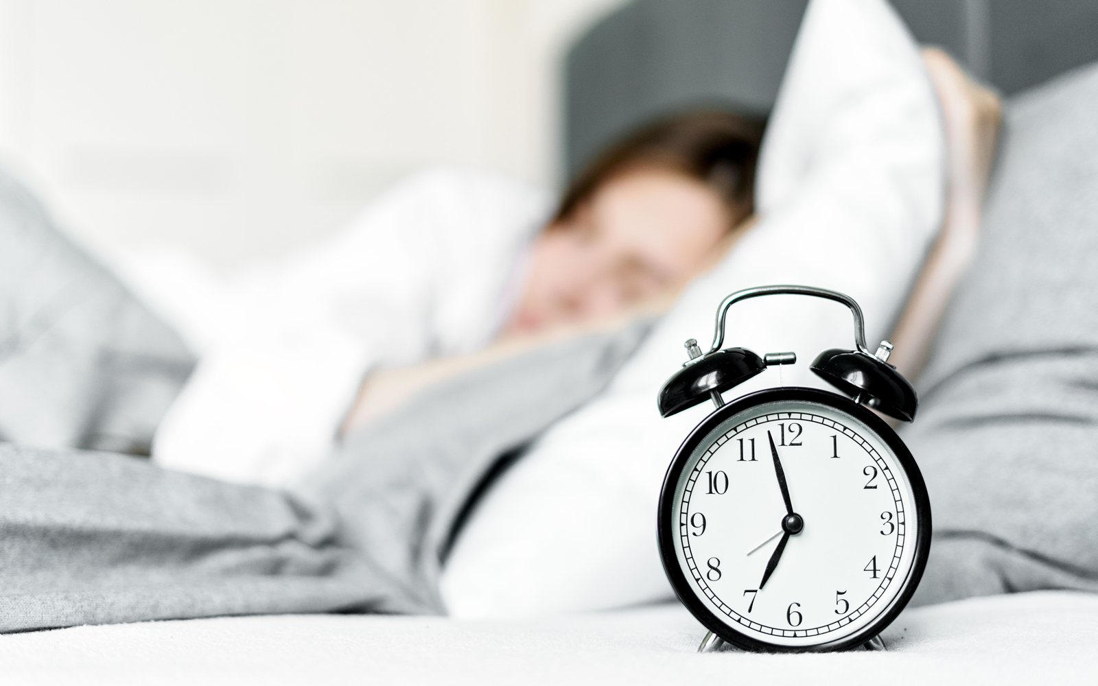 An alarm clock on a bed in the foreground with a woman asleep behind it